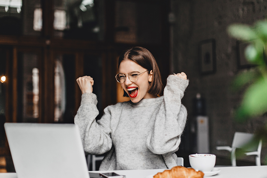 Happy woman in glasses makes winning gesture and sincerely rejoices. Lady with red lipstick dressed in gray sweater looking at laptop